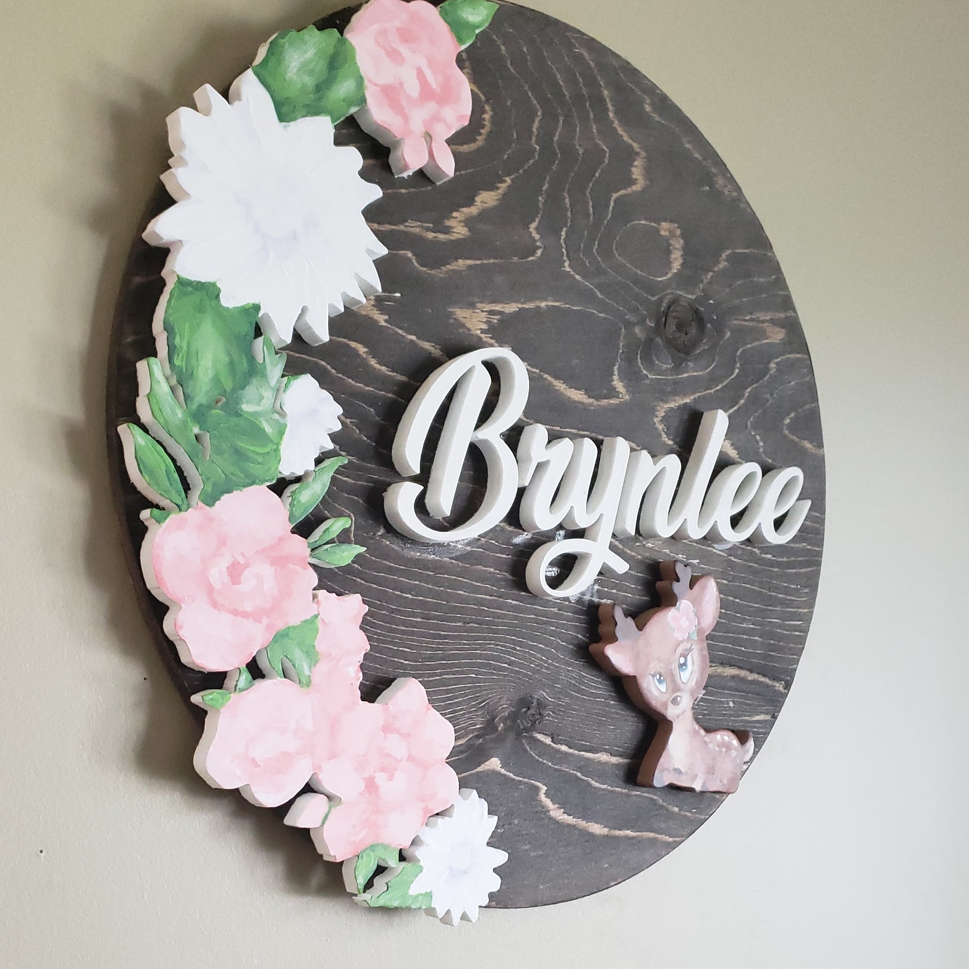 Espresso stained round sign, with the name Brynlee in white. Large mix of white, two toned pink flowers covers the left side of the sign with a deer sitting just beneath her name.