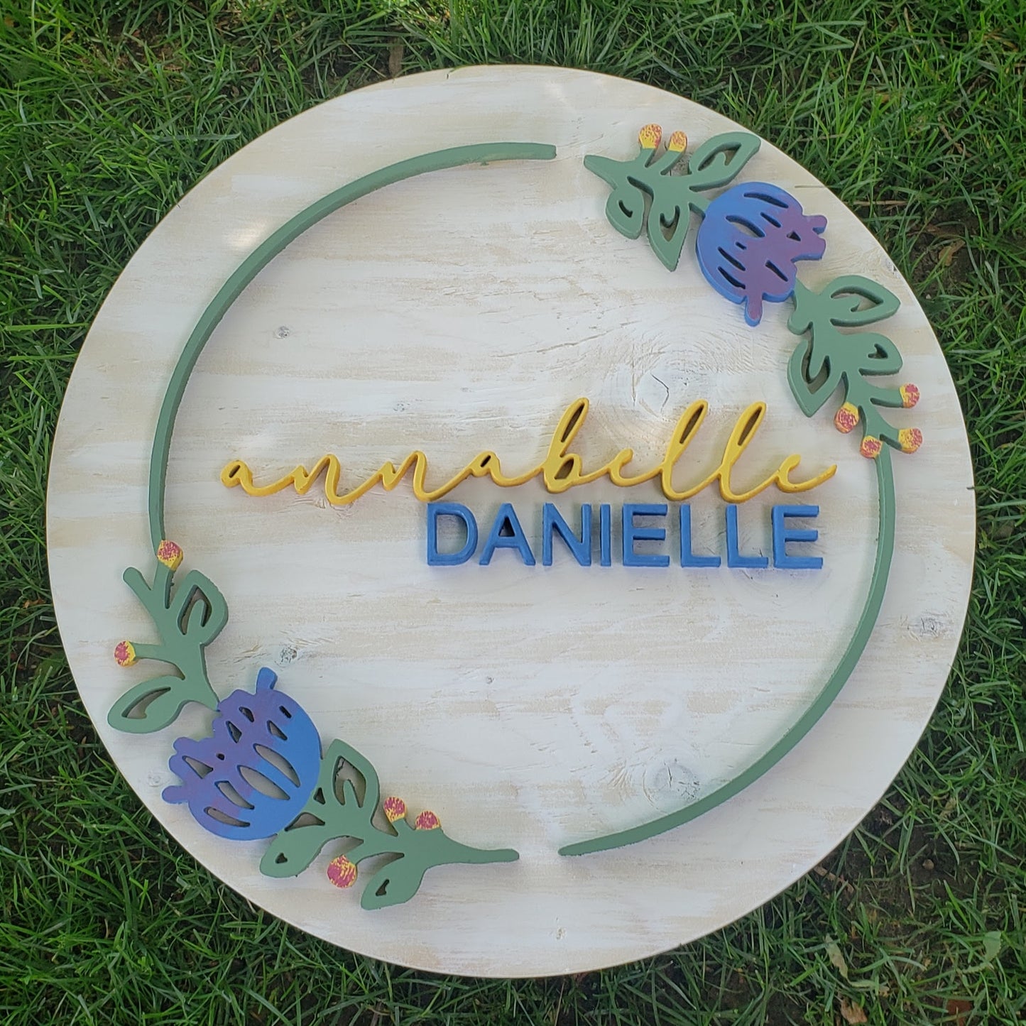 White washed round sign, with the name Annabelle Danielle surrounded by a simple elegant ring of greenery with purple flowers.