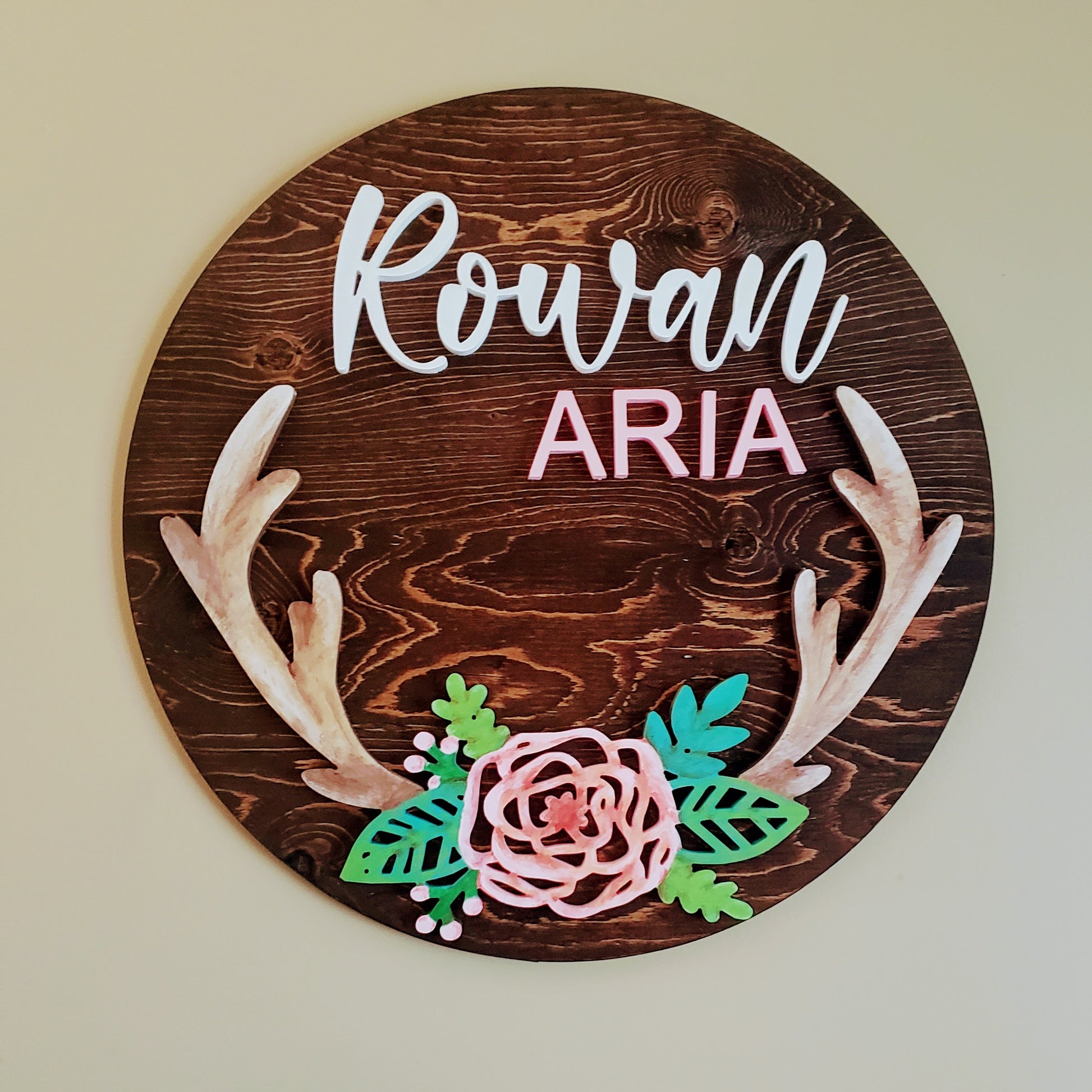 Medium wood stained round sign, with the name Rowan (in white) Aria (in pink), with pink flowers on the lower half some greenery and deer antlers coming out of the flowers 