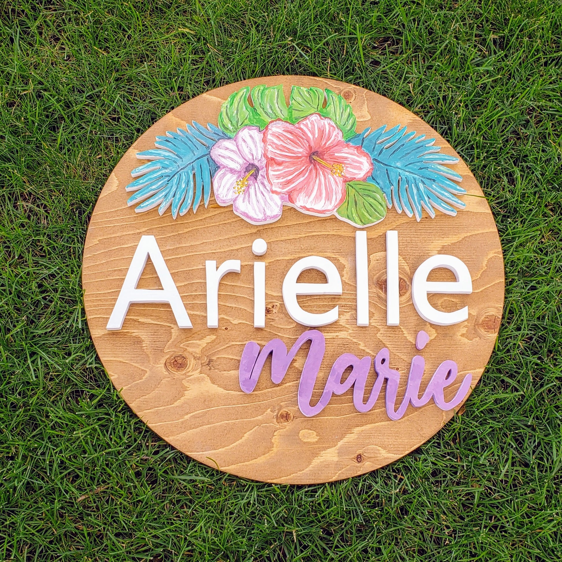 A round, pine stained sign, with the name Arielle Marie, accented with 2 hibiscus flowers (light and dark pink) and some palm leaves above.