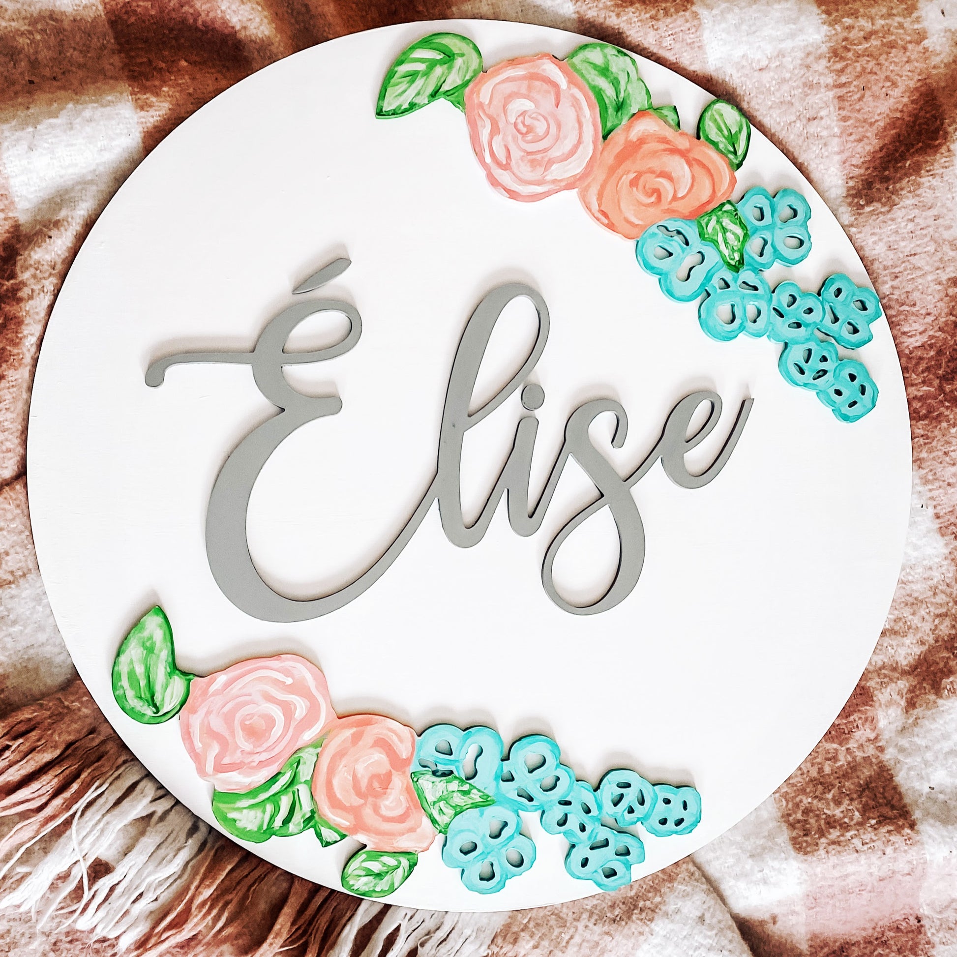 White round name sign, with the name élise in grey. Light and dark pink flowers, accompanied with blue flowers and greenery encases the name in a mirrored fashion