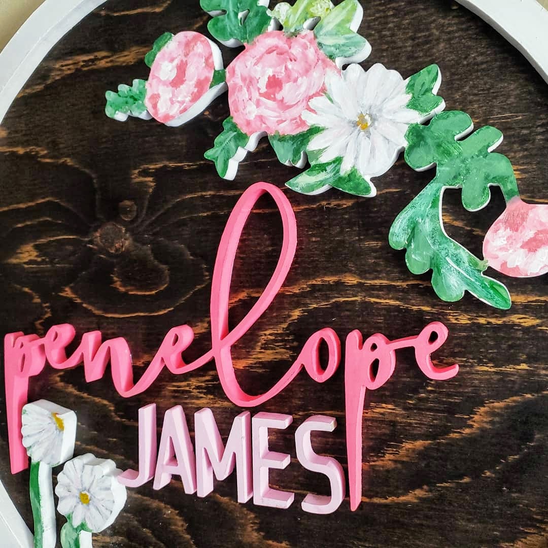 Espresso stained wood sign, with the name Penelope James, surrounded by white and pink flowers, enclosed in a white ring detail.