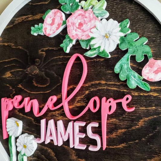 Espresso stained wood sign, with the name Penelope James, surrounded by white and pink flowers, enclosed in a white ring detail.