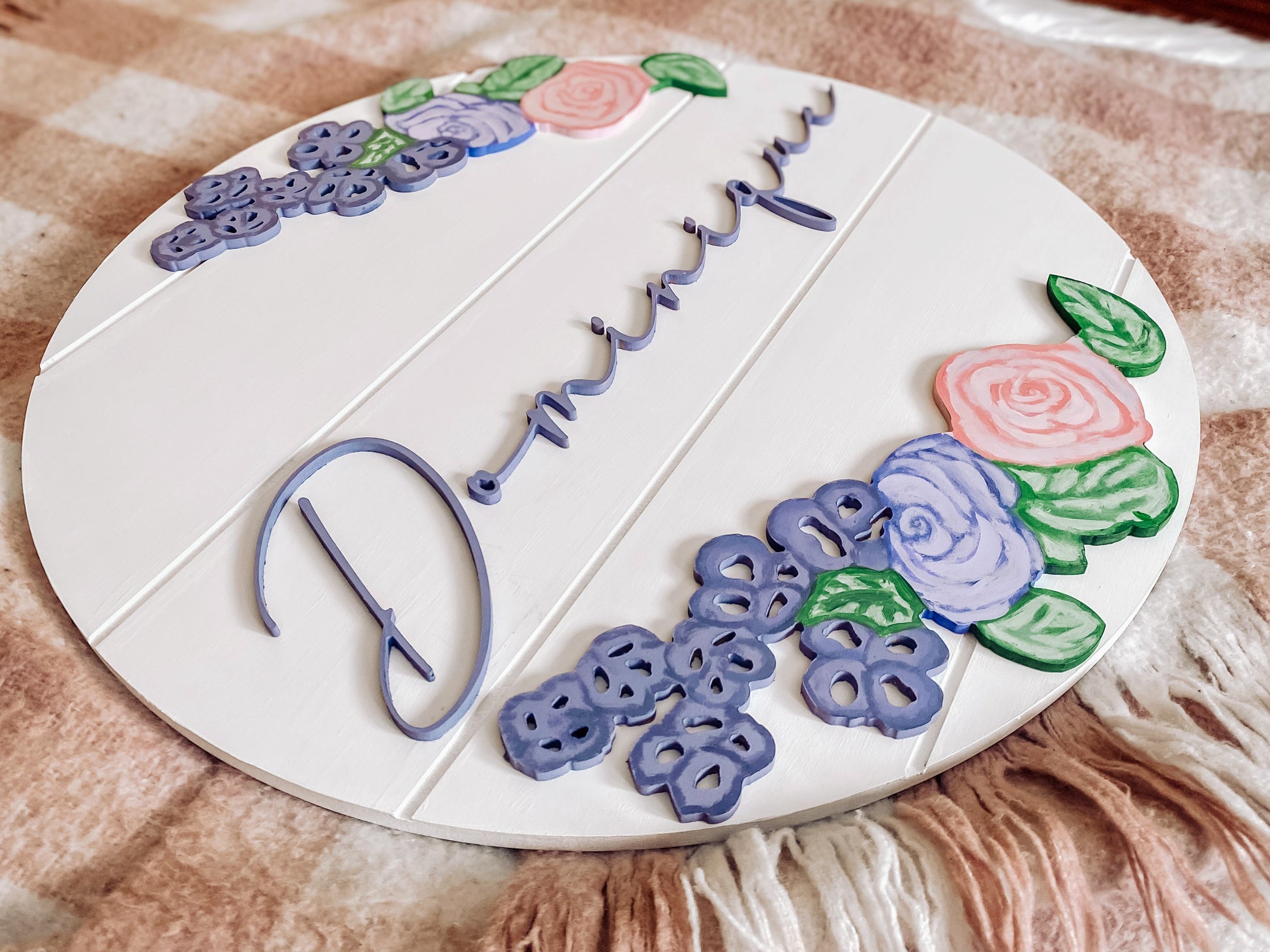 White shiplap round name sign, with the name Dominique in purple. Light pink and purple flowers, and greenery encases the name in a mirrored fashion