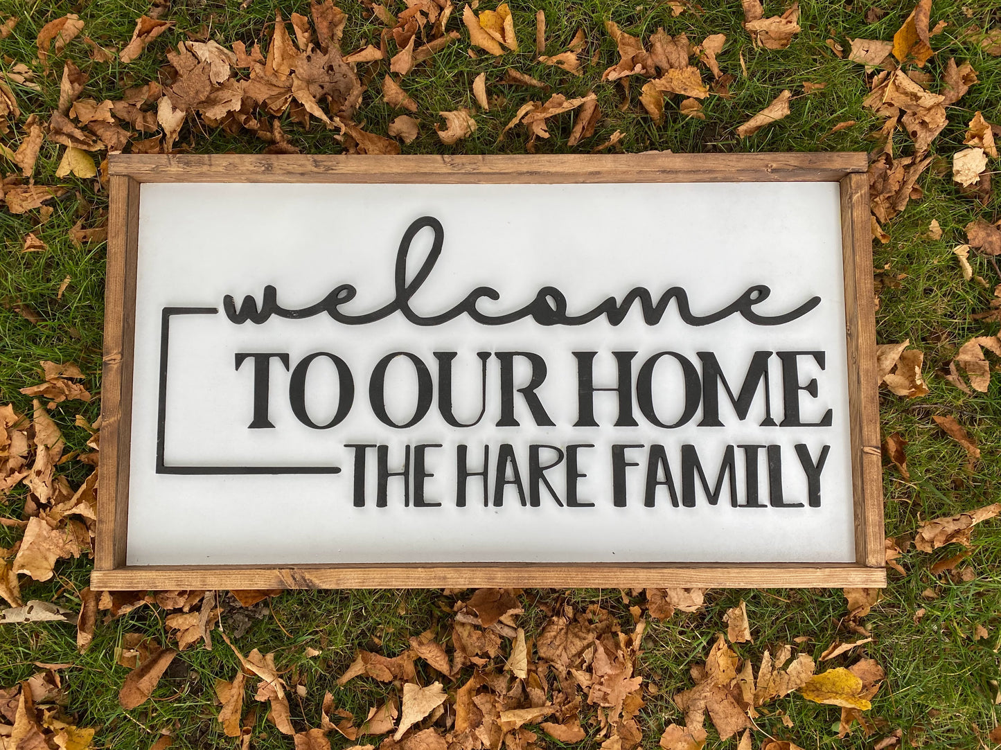 Rectangle sign with dark wood trim, white background and wording Welcome to our home, the hare family