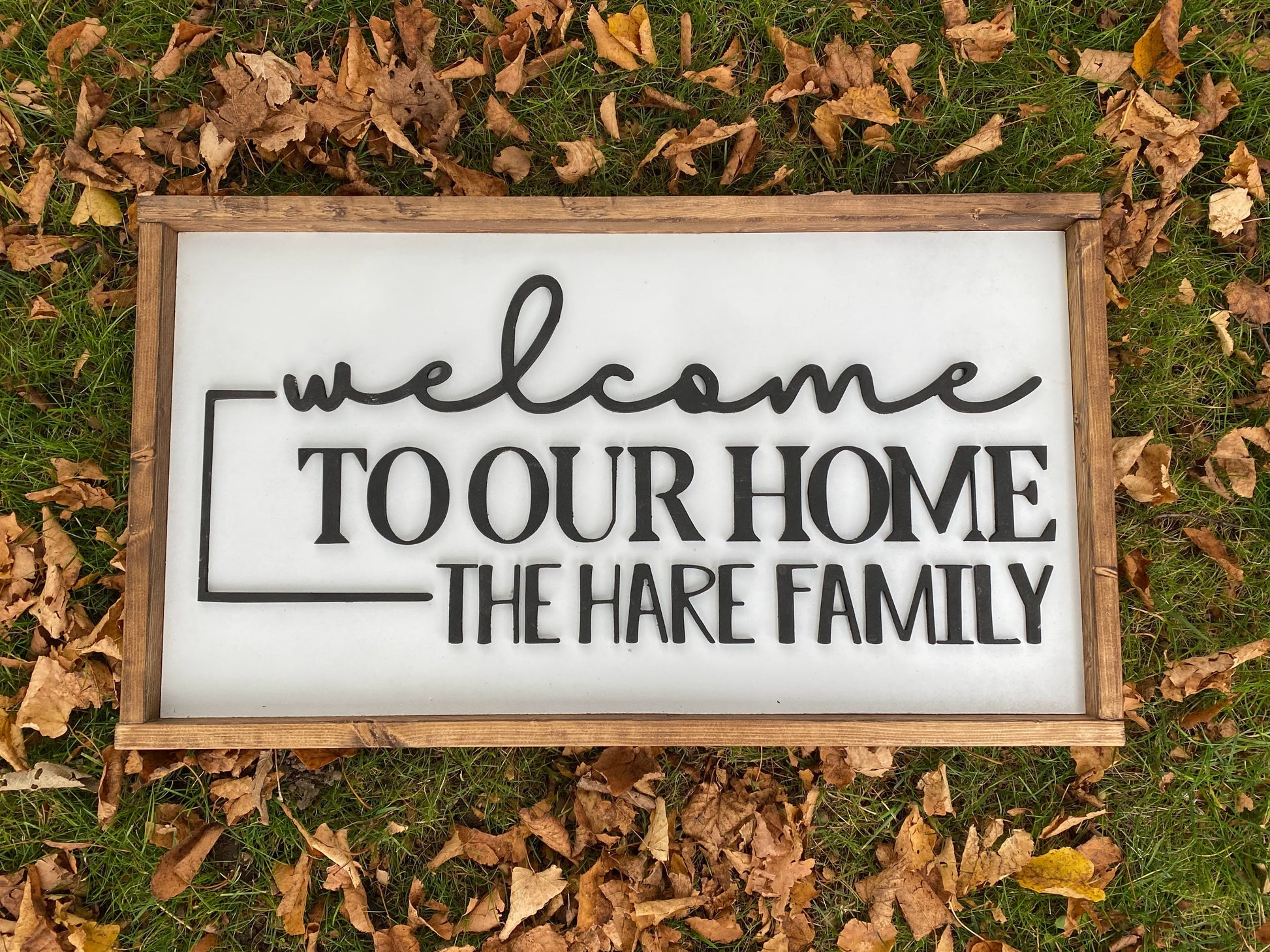 Rectangle sign with dark wood trim, white background and wording Welcome to our home, the hare family